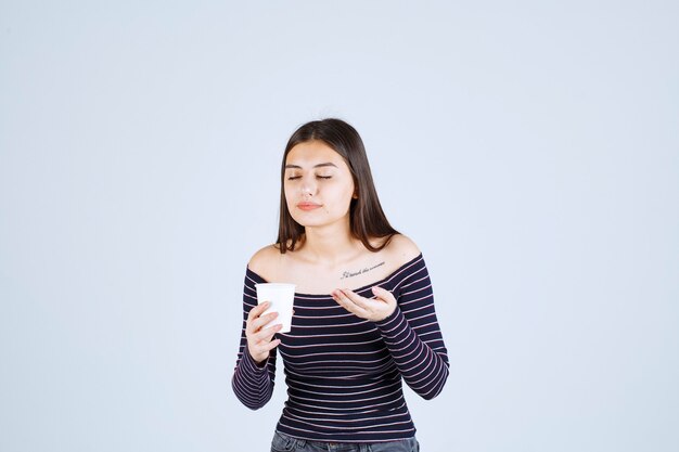 Chica en camisa a rayas sosteniendo una taza de café de plástico y oliéndola.