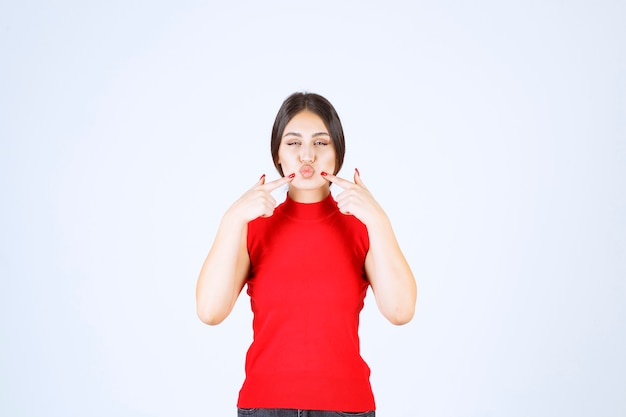 Chica de camisa roja apuntando a su boca y pidiendo silencio.