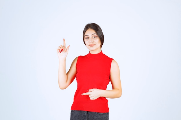 Chica de camisa roja levantando la mano y apuntando hacia arriba.