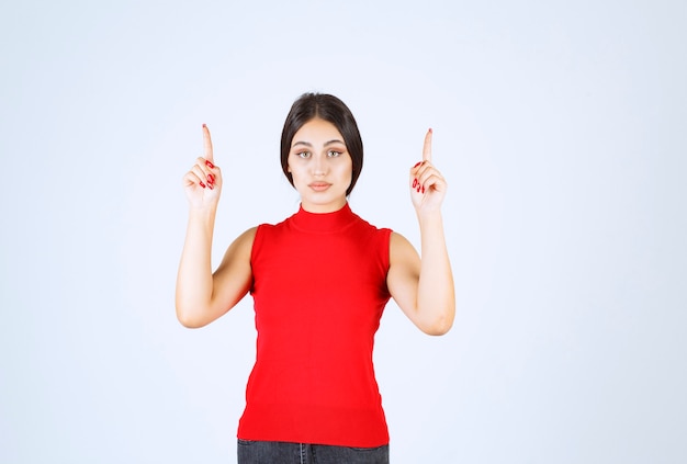 Chica de camisa roja levantando la mano y apuntando hacia arriba.