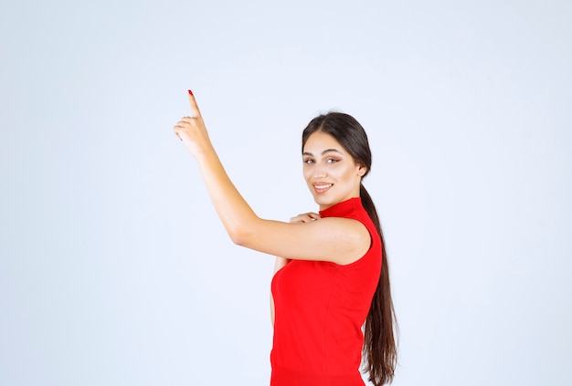 Chica de camisa roja levantando la mano y apuntando hacia arriba.