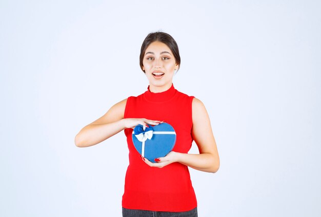 Chica en camisa roja presentando su caja de regalo con forma de corazón azul.