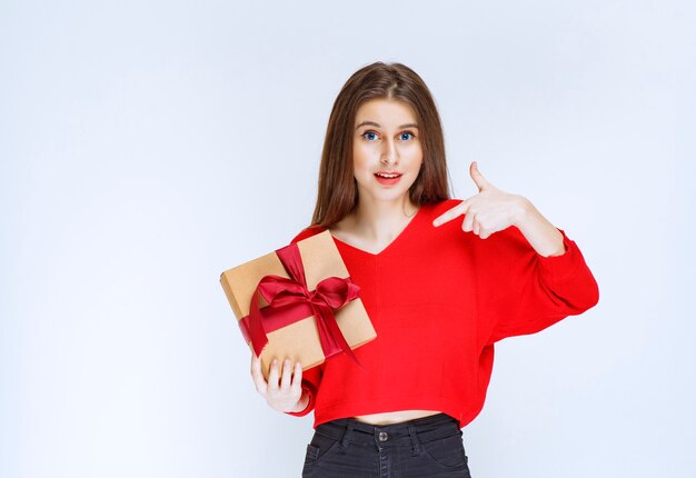 Chica en camisa roja sosteniendo una caja de regalo de cartón envuelta con cinta roja.