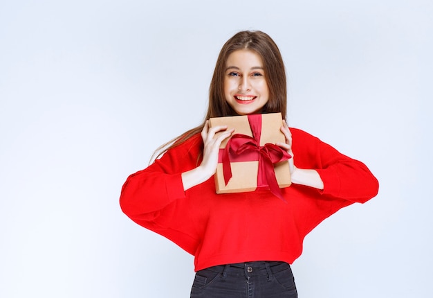 Chica en camisa roja sosteniendo una caja de regalo de cartón envuelta con cinta roja.