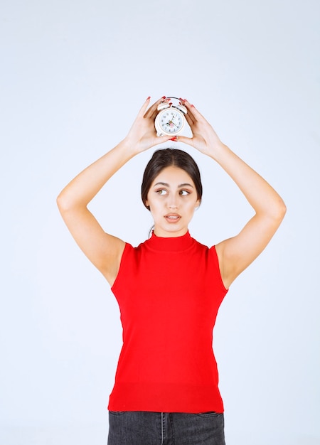 Chica en camisa roja sosteniendo un despertador sobre su cabeza.