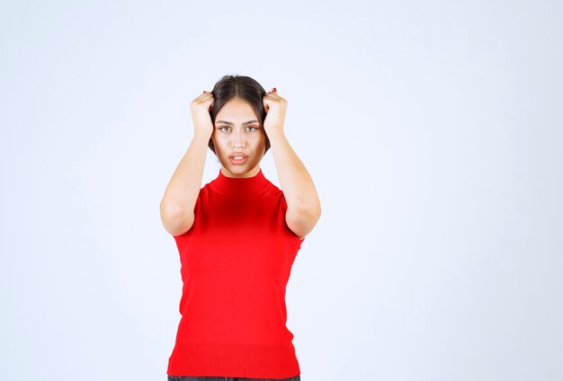 Chica de camisa roja sosteniendo su cabeza con las manos ya que tiene dolor de cabeza.