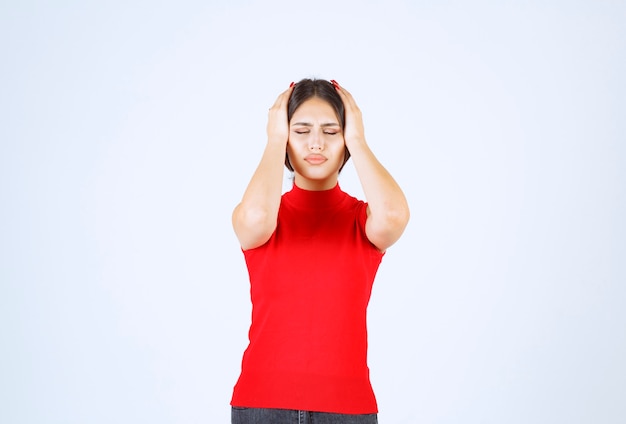 Chica de camisa roja sosteniendo su cabeza con las manos ya que tiene dolor de cabeza.