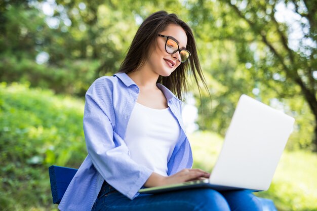 Chica en camiseta azul se sienta en el banco en el parque y usa su computadora portátil