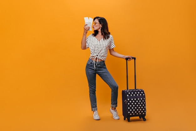 Chica en camiseta de lunares besa boletos y sostiene la maleta sobre fondo naranja