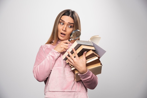 Chica en camiseta rosa sosteniendo una reserva de libros y tratando de leer el de arriba con una lupa.