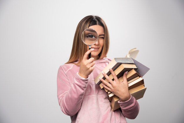 Chica en camiseta rosa sosteniendo una reserva de libros y tratando de leer el de arriba con una lupa.