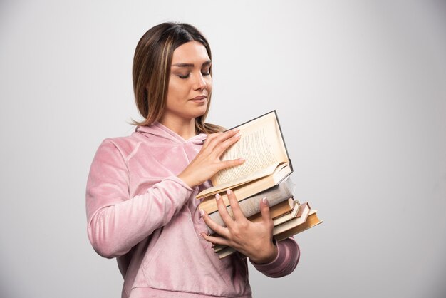 Chica en camiseta rosa sosteniendo un stock de libros, abriendo uno en la parte superior y leyéndolo