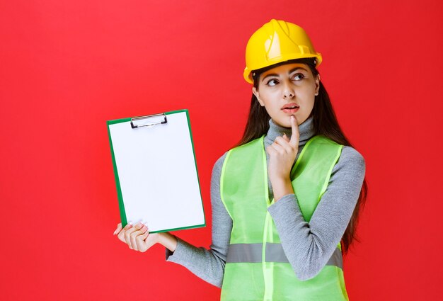 Chica de casco amarillo sosteniendo una carpeta de proyecto y pensando.