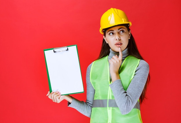 Chica de casco amarillo sosteniendo una carpeta de proyecto y pensando.