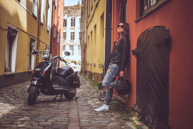 Chica casual con gafas de sol sosteniendo casco de moto negro y posando con moto scooter cerca de la vieja pared roja.