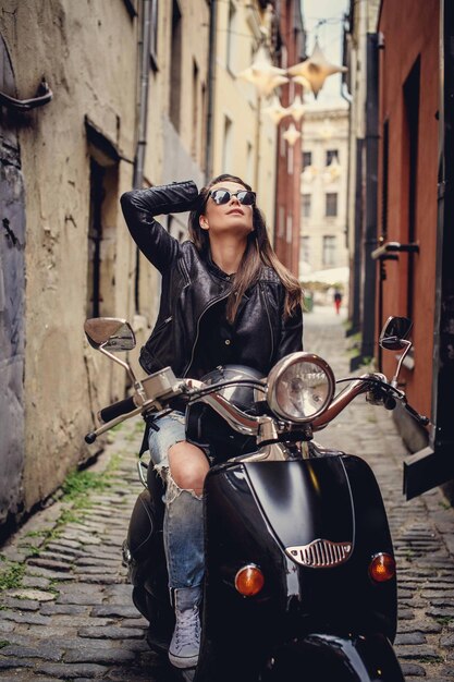 Chica casual en jeans y chaqueta de cuero posando en moto scooter en la calle del casco antiguo.