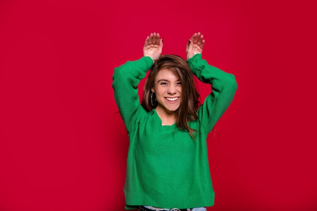 Chica caucásica alegre que expresa emociones positivas vestida con suéter verde mientras posa en año nuevo sobre fondo rojo con las manos sobre la cabeza. Mujer joven alegre en estudio