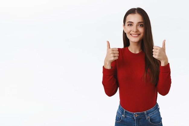 Una chica caucásica bastante amistosa y solidaria con suéter rojo como idea para organizar una fiesta de reunión familiar de acción de gracias mostrando el pulgar hacia arriba en aprobación o aceptación sonriendo y asintiendo de acuerdo