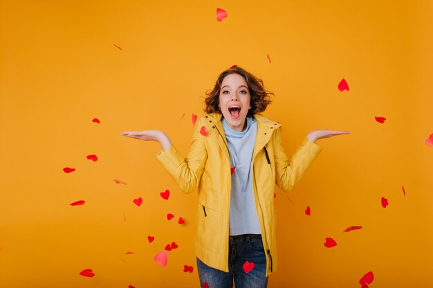 Chica caucásica complacida en elegante chaqueta amarilla que expresa felicidad en el día de San Valentín. Modelo de mujer alegre con cabello ondulado tirando corazones.