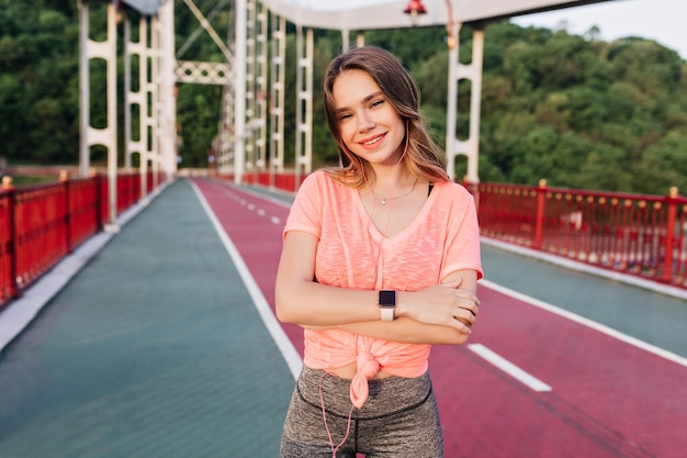 Chica caucásica complacida con smartwatch posando en camino de ceniza. Tiro al aire libre de la alegre dama rubia de pie en el estadio con una sonrisa suave.