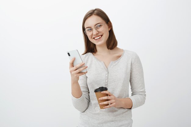 Chica caucásica entusiasta de aspecto amistoso con gafas con cabello corto castaño sosteniendo smarpthone y taza de café de papel esperando amigo en la cafetería y jugando en la aplicación usando un nuevo teléfono celular sobre una pared gris