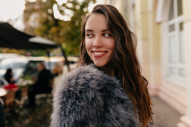 Chica caucásica con estilo atractiva en abrigo de piel mirando a otro lado y sonriendo en el fondo de la ciudad.