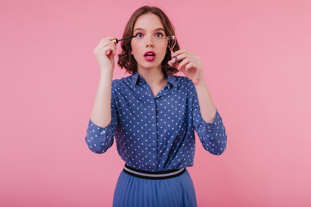 Chica caucásica con estilo haciendo maquillaje de ojos. Foto interior de mujer morena divertida riza sus pestañas en la pared rosa.