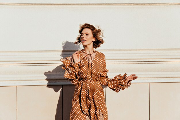 Chica caucásica extática con pelo corto oscuro mirando a su alrededor. Foto al aire libre de adorable mujer blanca con elegante vestido marrón.