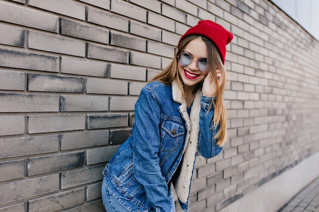 Chica caucásica extática en traje de mezclilla y gafas azules posando con una linda sonrisa. Mujer joven complacida con sombrero rojo jugando durante la sesión de fotos en la calle.