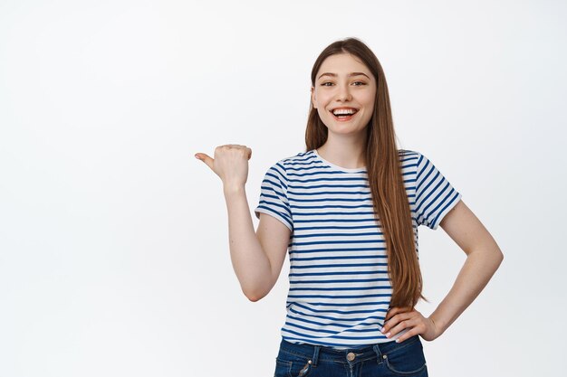 Chica caucásica feliz riendo, sonriendo y señalando casualmente con el dedo a la izquierda en el espacio vacío, banner de venta de compras, de pie sobre fondo blanco.