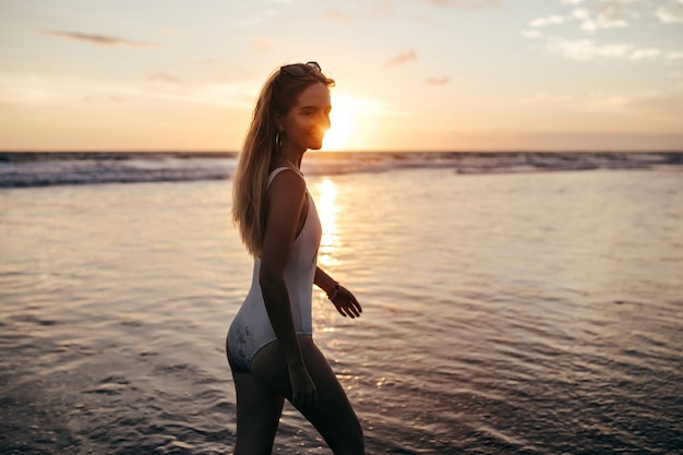 Chica caucásica inspirada en traje de baño blanco divirtiéndose en el mar por la noche. Fascinante joven posando en el océano con puesta de sol