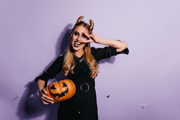 Chica caucásica inspirada en traje mágico posando en la pared púrpura. Vampiro mujer elegante con calabaza de halloween con sonrisa.