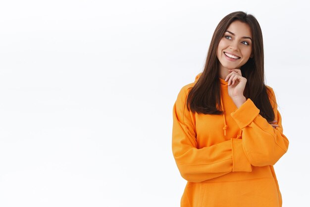 Chica caucásica intrigada y pensativa con capucha naranja toca la barbilla inclina la cabeza y mira a la izquierda curiosa sonriendo interesada contemplar cosas geniales haciendo elección de pie fondo blanco reflexionar