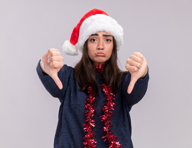 Chica caucásica joven decepcionada con gorro de Papá Noel y guirnalda alrededor del cuello pulgares hacia abajo