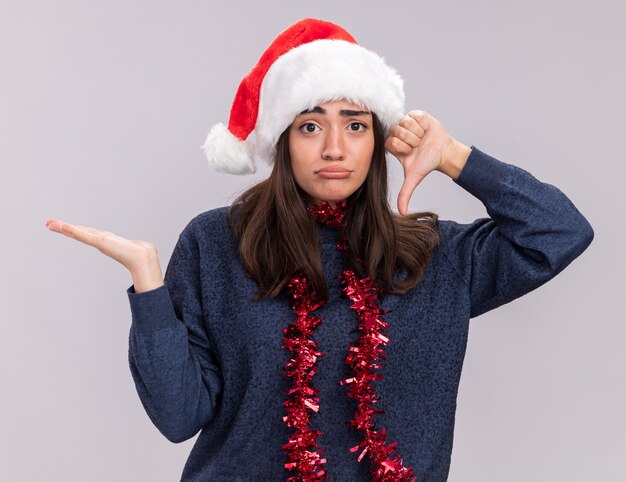 Chica caucásica joven decepcionada con gorro de Papá Noel y guirnalda alrededor del cuello sostiene la mano abierta y el pulgar hacia abajo aislado en la pared blanca con espacio de copia