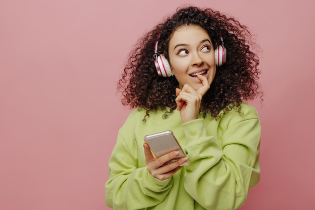 Chica caucásica joven despreocupada se pone los auriculares conecta el teléfono inteligente elige la canción en el interior Morena en suéter verde mira hacia arriba Concepto de tecnología y estilo de vida