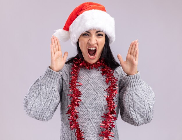 Chica caucásica joven furiosa con sombrero de navidad y guirnalda de oropel alrededor del cuello manteniendo las manos en el aire gritando aislado en la pared blanca