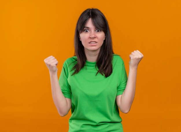 Chica caucásica joven molesta en camisa verde levantó los puños y mira a la cámara sobre fondo naranja aislado