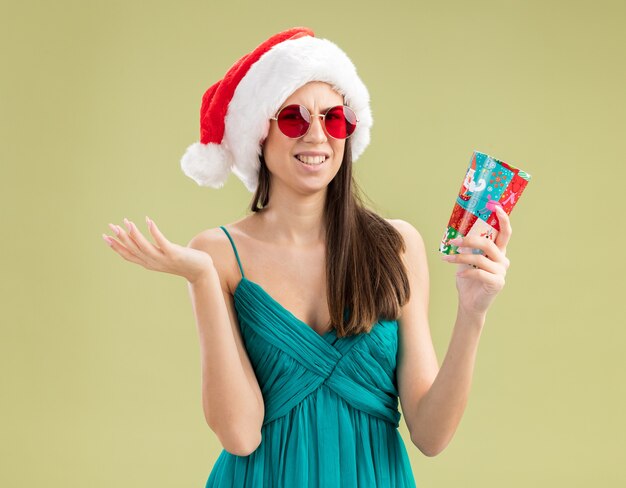 Chica caucásica joven molesta en gafas de sol con sombrero de santa sosteniendo una taza de papel