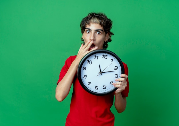 Chica caucásica joven sorprendida con corte de pelo pixie sosteniendo el reloj con la mano en la boca aislada sobre fondo verde con espacio de copia