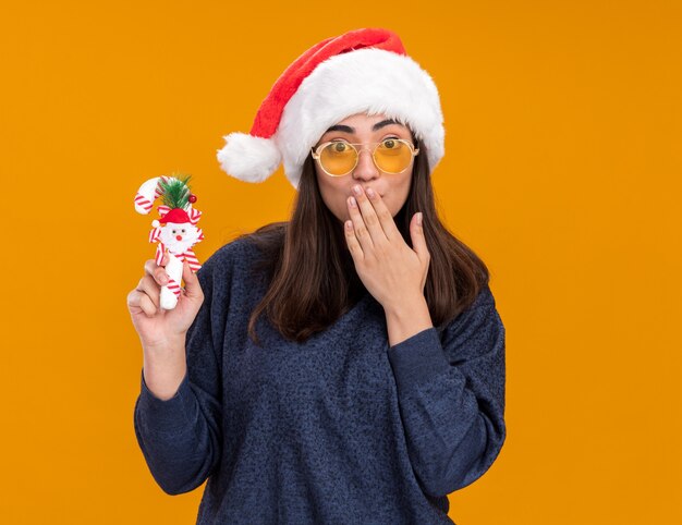 Chica caucásica joven sorprendida en gafas de sol con sombrero de santa sostiene bastón de caramelo y pone la mano en la boca aislada en la pared naranja con espacio de copia