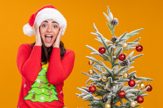 Chica caucásica joven sorprendida con sombrero de santa pone las manos en la cara de pie junto al árbol de Navidad aislado sobre fondo naranja con espacio de copia