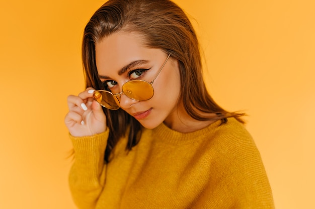 Chica caucásica de moda mirando a través de gafas de sol amarillas. Modelo de mujer dichosa en elegante traje de otoño posando.