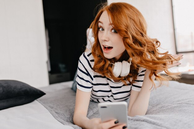 Chica caucásica de moda posando en la cama con sonrisa de sorpresa. Modelo de jengibre alegre en auriculares blancos en dormitorio.