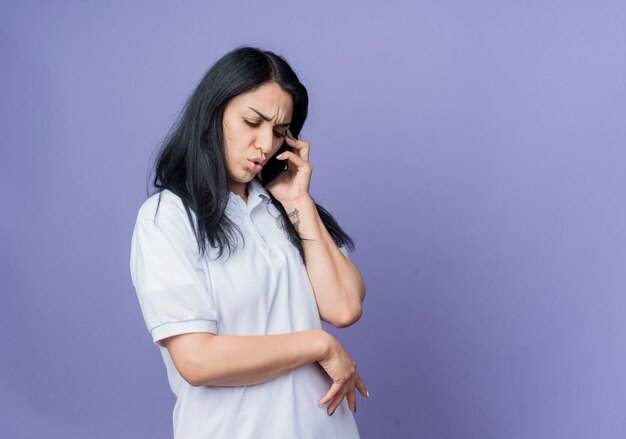 Chica caucásica morena joven disgustada habla por teléfono mirando hacia abajo aislado en la pared púrpura