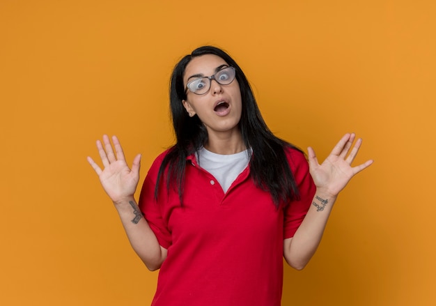 Chica caucásica morena joven sorprendida en gafas ópticas con camisa roja levanta las manos mirando aislado en la pared naranja