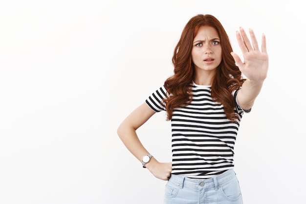 Chica caucásica pelirroja mandona de aspecto serio con camiseta a rayas tirar de la mano en stop motion, prohibir un paso más allá, dar prohibición, evitar que un amigo beba alcohol, pared blanca