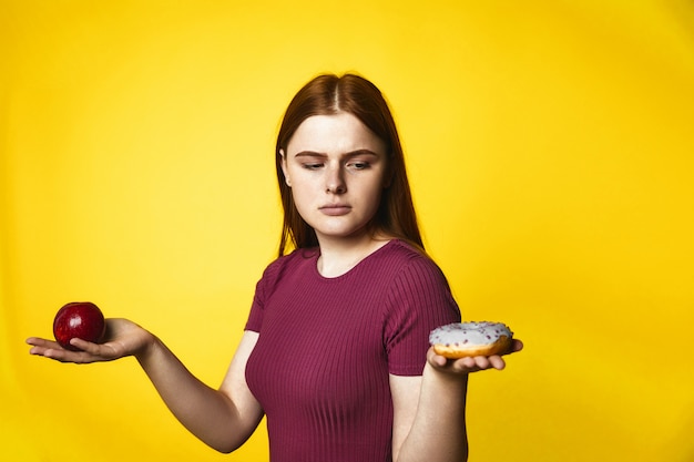 Chica caucásica pelirroja pensativa sostiene manzana en una mano y donut en otra