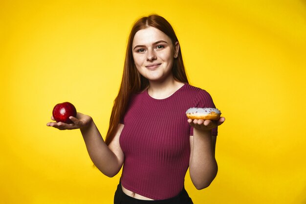 La chica caucásica pelirroja sonrió sostiene la manzana en una mano y la rosquilla en la otra