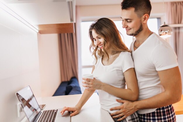 Chica caucásica de pelo oscuro bebiendo té mientras se relaja con su marido en la mañana. Feliz pareja europea usando laptop en fin de semana en casa.